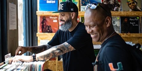 A man with tattooed arms wearing black cap flipping through a stack of vinyl records and another man with eyeglasses raised on his head